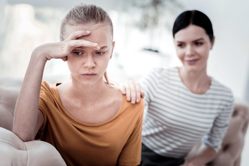 Do not worry. Portrait of sad girl keeping her hand on her forehead and looking down while her mother calming her down - obrazy, fototapety, plakaty