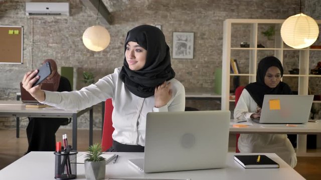 Three young muslim womens in hijab sitting and typing on laptop in modern office, charming muslim women taking selfie with phone