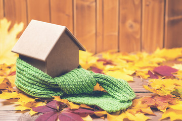 Green scarf around a miniature house on autumn leaves background. Concept of protecting or...