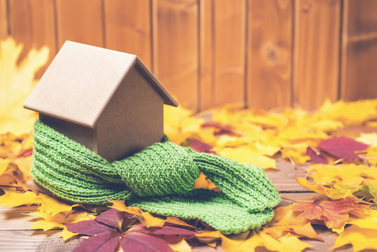 Green scarf around a miniature house on autumn leaves background. Concept of protecting or isolating house.