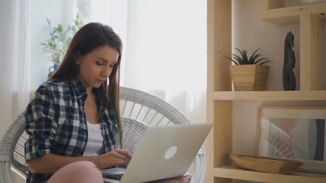 Young woman freelancer is working with laptop sitting in apartment, beautiful female is typing text, looking at device screen, performing multitasking job in armchair at home. Concept: home work