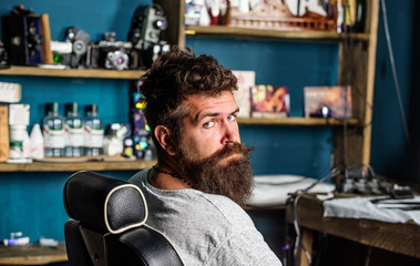 Man with beard and mustache sits in hairdressers chair, beauty supplies on background. Hipster with...