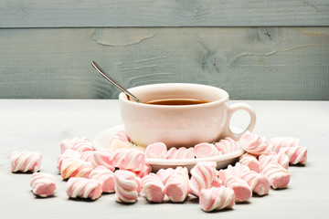 Cup with hot black tea and marshmallows. Cup and sweets prepared as dessert. Tea break concept. Mug filled with black brewed tea, spoon and heap of marshmallow on white wooden background