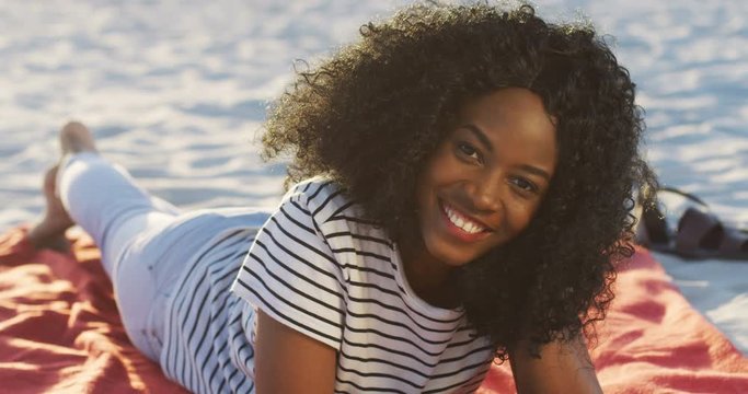 Close up of the young pretty curly African American womanlying on the sea beach and smiling to the camera. Outside.