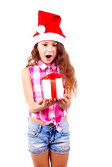 Cute little girl in santa hat holds a gift in her hand and opens her mouth in surprise