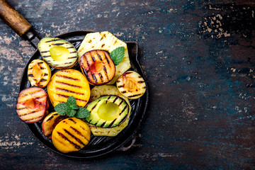 GRILLED FRUITS. Grill fruits - pineapple, peaches, plums, avocado, pear on black cast iron grill pan. Dark background. Top view. Copy space