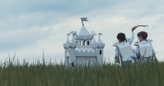 Brothers boys wearing cardboard medieval knight armor costumes riding stick horses through a grass field in summer. 4K UHD 60 FPS SLO MO