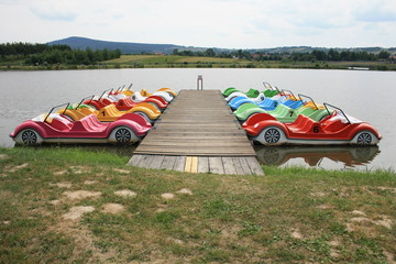 Rowery wodne w kształcie samochodów zacumowane do drewnianego pomostu nad jeziorem w Wilkowie, świętokrzyskie