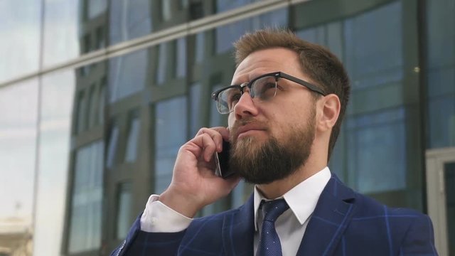 Portrait of solid young man talking on phone standing on modern building backgroung, bearded businessman wearing glasses is makes call, discussing startup project on street in summer day. Concept