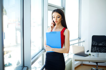 Naklejka premium Business. Beautiful Young Woman Works in Light Office. Dressed in Red T-shirt and Black Skirt. Girl Sitting on Tables Holds Folder Hand and Smiles. High Resolution