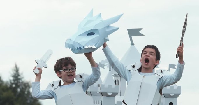 Brothers kid boys wearing medieval knight armor costume celebrating, holding a dragon head mask near cardboard castle. 4K UHD 60 FPS SLO MO