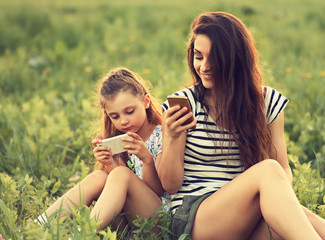 Naklejka premium Happy smiling mother and fun kid girl sitting on green glass and playing in mobile phone. Contemporary family lifestyle. Concept. Toned closeup portrait