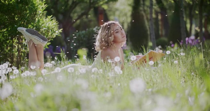 beautiful Asian girl surrounded by dandelions in the Park on a Sunny day with a book, having fun slow motion