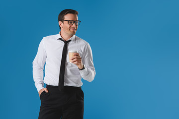 portrait of handsome smiling businesman in eyeglasses holding paper cup and looking away isolated on blue