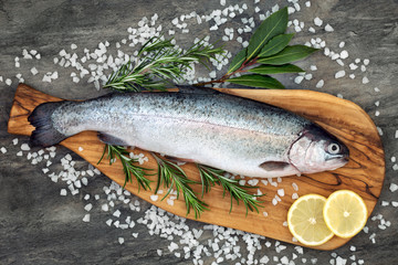 Rainbow trout healthy heart food on an olive wood board, with rosemary and bay leaf herbs, course sea salt and lemon on marble background. High in omega 3 fatty acid. - obrazy, fototapety, plakaty