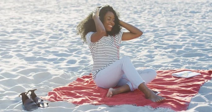 Beautiful curly African American young woman in the headphones listening to the music on the smartphone and dancing while sitting on the beach. Outdoor.