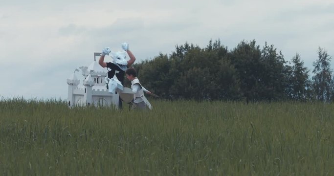 Kid boy wearing medieval knight armor costume fighting his father pretending to be a dragon in paper mask near cardboard castle. 4K UHD 60 FPS SLO MO