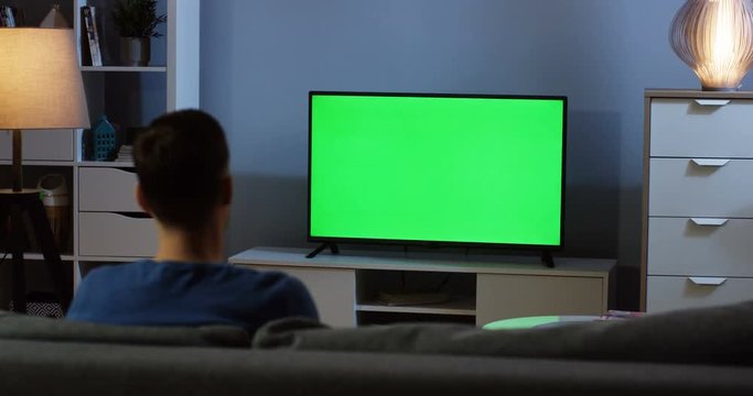 Rear view on the young man sitting on the sofa in the living room, watching TV with green screen and changing channels with a remote control in the evening. Chroma key. Inside