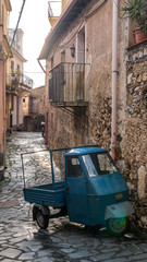 Vintage vehicle with three weels and blue metal paint, called Ape car,  parked on the street, next to a rural house with big stone wall. - Castelmola, Taormina, Messina. March 2017