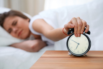 Sleepy young woman stretching hand to ringing alarm willing turn it off. Early wake up, not getting enough sleep concept