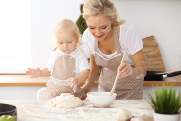 Little girl and her blonde mom in beige aprons  playing and laughing while kneading the dough in the kitchen. Homemade pastry for bread, pizza or bake cookies