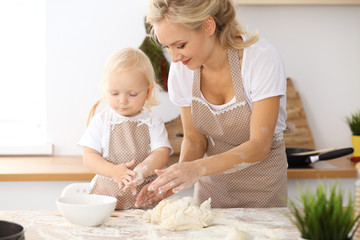 Little girl and her blonde mom in beige aprons  playing and laughing while kneading the dough in the kitchen. Homemade pastry for bread, pizza or bake cookies