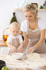 Little girl and her blonde mom in beige aprons  playing and laughing while kneading the dough in the kitchen. Homemade pastry for bread, pizza or bake cookies