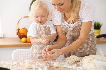 Little girl and her blonde mom in beige aprons  playing and laughing while kneading the dough in the kitchen. Homemade pastry for bread, pizza or bake cookies