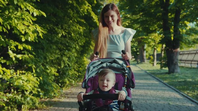 Beautiful young woman in a light summer dress pushes her cute son in a baby stroller down the alley, smiles, talks to him. Family bonds, cheerful mood, summertime. Happy childhood. Family portrait