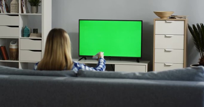 Rear view on the blonde woman in the plaid shirt sitting on the gray sofa in the living room, watching TV with green screen and changing channels with a remote control. Chroma key. Indoor