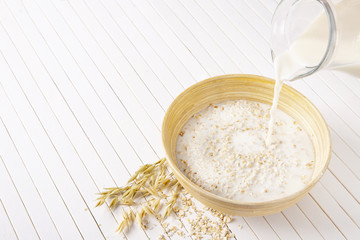 Oatmeal flakes in a wooden bowl on a white background. Milk pours into a bowl of cereal.