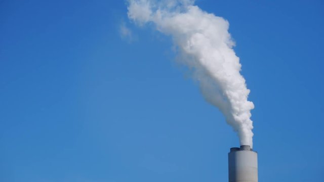 A close up shot of white smoke billowing from a tall smokestack on a factory on a sunny summer day. Shot at 60fps for optional slow motion usage.  	