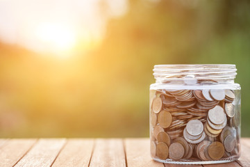 Thai coins in money jar on wooden plank wirh sublight and blur background. Savings concept.