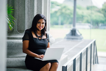 An Indian professional woman sits on a concrete bench during the day and is working on her laptop. She is smiling confidently as she continues her work.