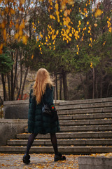 Red-haired girl in autumn park. She's wearing shorts and gloves and with bag. Woman is walking. Man is unrecognizable.