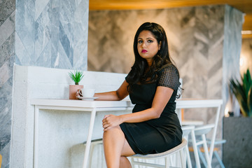 A successful Indian woman leader looking serious while having her coffee break in a cafeteria. She is looking very fierce and elegant on her classy black dress.