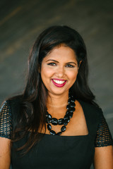 A studio portrait of a youthful but confident and successful Indian woman in a studio. She is dressed in an elegant and professional black dress and is smiling for her professional head shot.