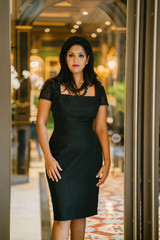 A confident desi woman stands across the hallway of their business office. She looks dignified in her black dress.