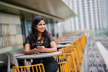 A confident Indian Asian woman is sending an SMS on her smartphone during the day. She is sitting outdoors in a row of trendy yellow chairs.