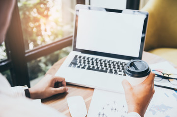 Business man hands holding cup of  coffee  and using laptop with blank screen on desk in cafe.