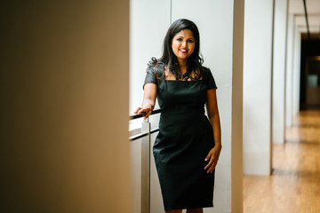 An Indian Asian female professional is standing in the corridor of a corporate building during the day. She is very elegant and is smiling with confidence.