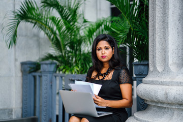 A mature and confident Indian Asian woman sits on the steps of a legal looking building and is working on her laptop computer with some documents. 