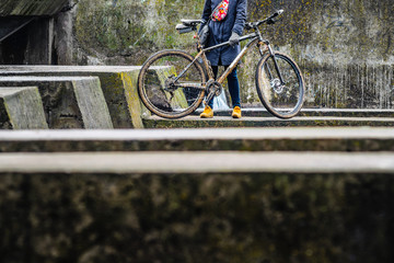 Figure of a Man Next to a Dirty Mountain Bike Against the Background of Old Urban Concrete Structures. Dirty Mountain Bike Covered with Mud After Riding in Bad Weather Stands. Grey 29er Hardtail Bike