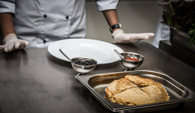 Pancakes and two kinds of caviar in metal dishes on a professional restaurant kitchen. Hands of Chef. Selective focus. Shallow depth of field. Toned