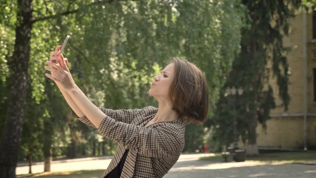 Young attractive girl is making selfie in park in daytime, duck face, in summer, communication concept, side view