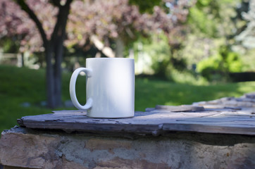 A blank white coffee mug on the ledge of a park under the pink trees. 