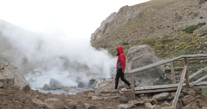 Detail of blasting Geysers, Los tachos, in rocky coast of Covunco warm river. Woman with jacket walks over bridge and stands to see the geysers. Background of valley and snowy mountains.