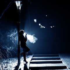 Portrait of a smoking woman in the style of noir. A woman with a cigarette is standing by the street lamp at night.