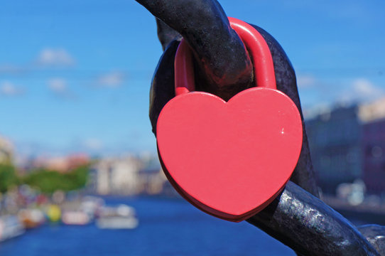 A red heart shaped lock connected to chain, canal with boats in the background.              