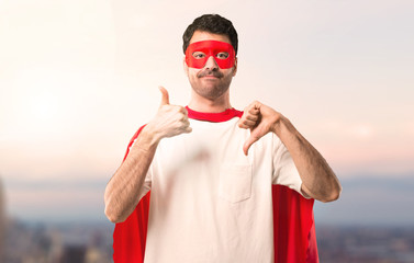 Naklejka premium Superhero man with mask and red cape making good-bad sign. Undecided person between yes or not on a sunset background